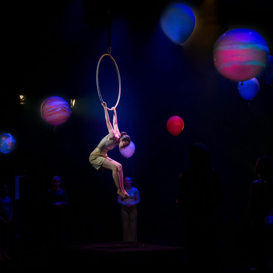 Akrobatin schwingt an einem Ring, umgeben von bunten Luftballons und gedämpftem Licht.